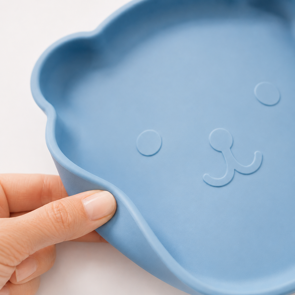 Macro del piatto in silicone blu a forma di orsetto per lo svezzamento, con superficie opaca e flessibile evidenziata da una mano che piega delicatamente il bordo.